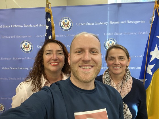 Ela, Haris, and Selma of USAID/Bosnia and Herzegovina smile for a selfie in front of a blue United States Embassy, Bosnia and Herzegovina backdrop.