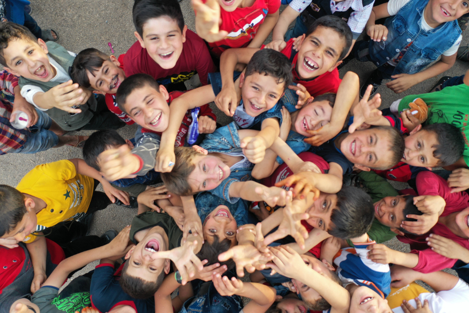 Over head photo of children looking up at camera above. They are standing in a circle smiling. 