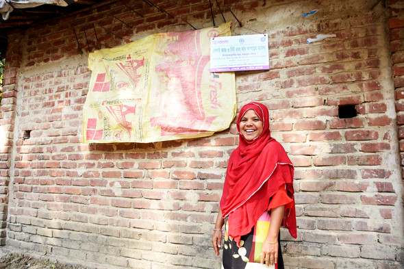 Mahamuda Begum poses outside of her brick home, smiling at the camera.