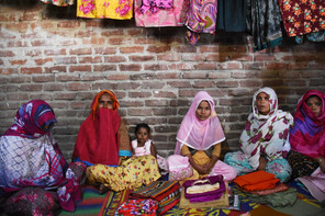 Female community members sit on the ground in Mme. Begum's home surrounded by bright fabric.