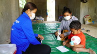 A young child with a soccer ball in hand is sitting on the ground writing while a social worker watches. 