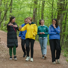 A group of five people walk on a tree-lined trail as one of them talks and gestures with her hands.