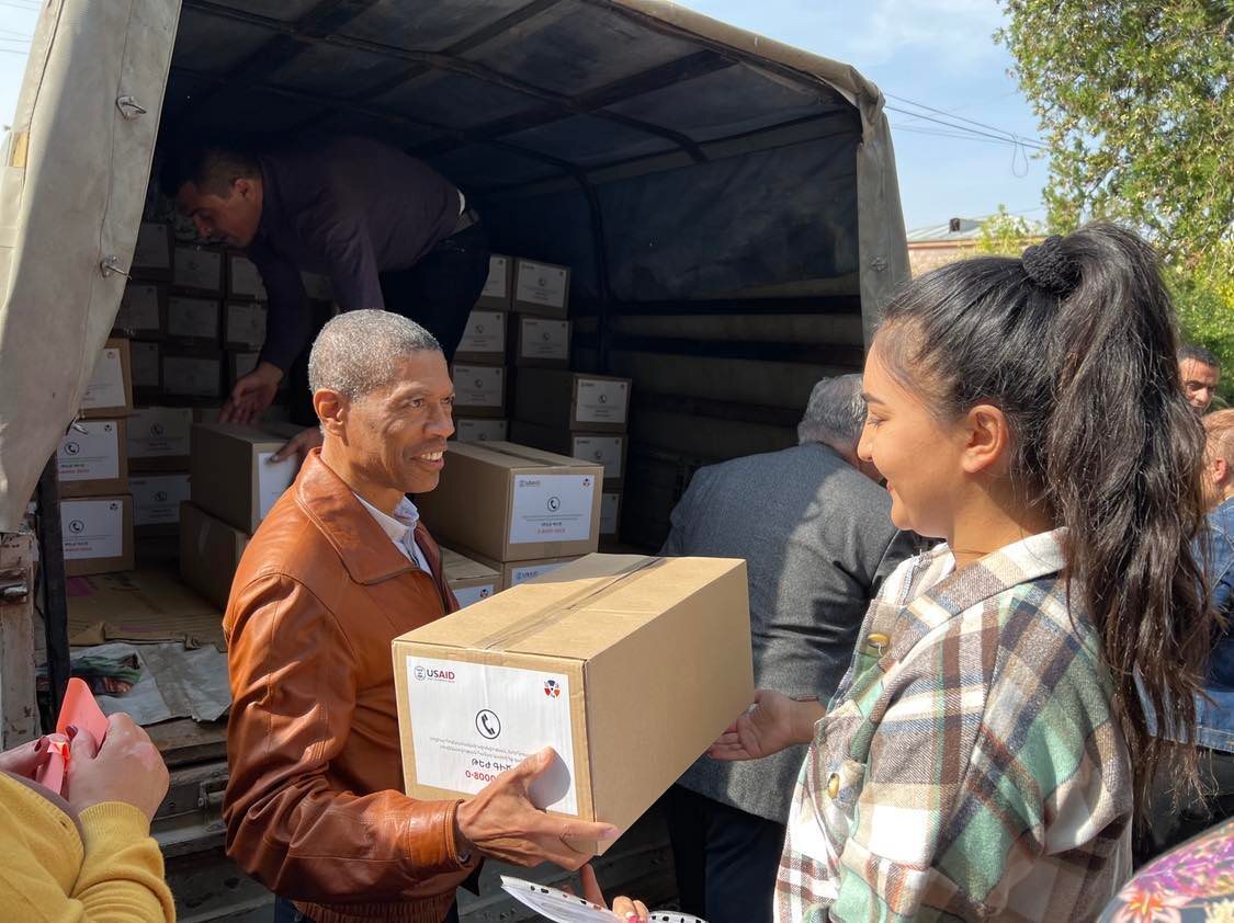 By an open truck filled with supply boxes, a smiling man passes a box marked with USAID and its partner’s logos to a woman.