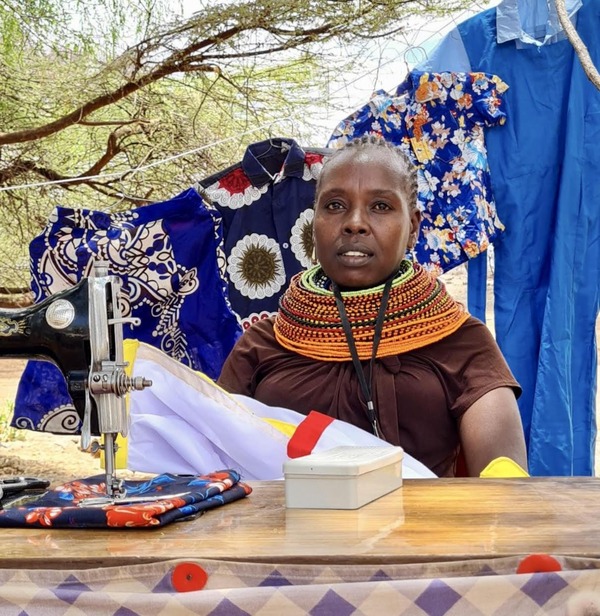 Jane sits at a table with her sewing machine with brightly colored fabrics hanging behind her.