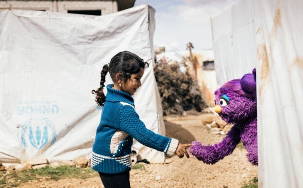 A young girl touches hands with a purple stuffed animal from Sesame Street at a refugee camp.