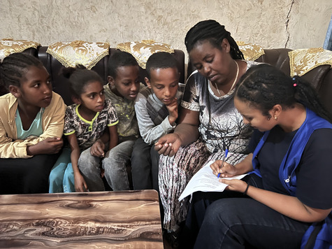 Four young children and an older woman look at a piece of paper as a fifth woman in a blue vest writes on it with a blue pen.