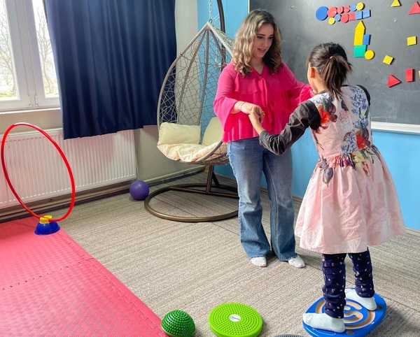 A woman holds a young girl's hands, helping her with a fun balance activity in a colorful room designed for youth.