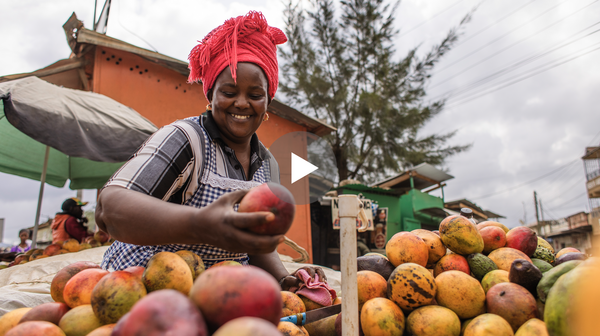 Video still of a woman with a red head wrap picking fruit from a pile