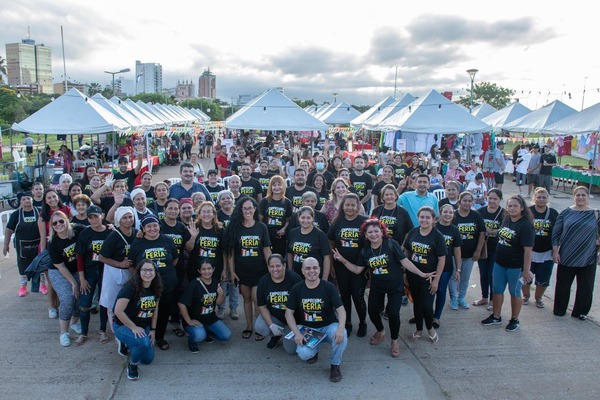  A group of more than 50 individuals wearing matching shirts pose in front of white canopy tents at an outdoor market.