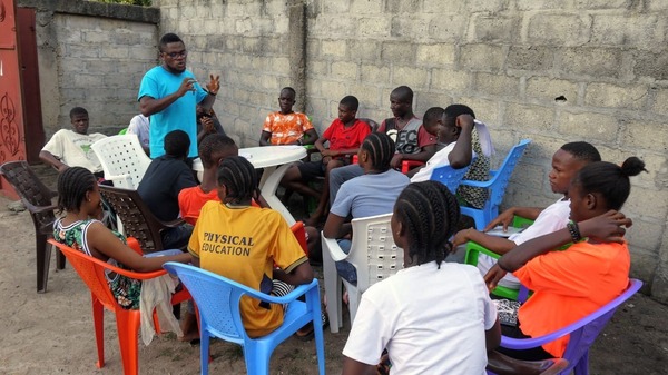 A man speaks to a group of 16 youth in a circle in plastic chairs in a courtyard with walls of cement blocks and a dirt floor.