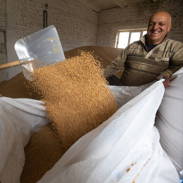 A smiling man holds a large white bag being filled with grain