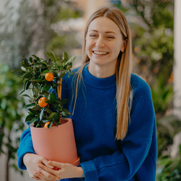 A smiling woman with blonde hair and a blue sweater holding a green plant with oranges