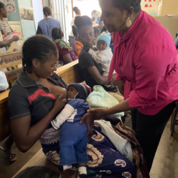 Nutritionist wearing a bright pink blouse gently helps a mother position her son to latch onto her breast.