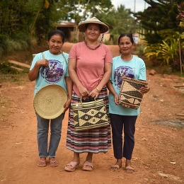 A woman wearing a pink shirt and straw hat stands in between two women in teal shirts. All three hold woven straw items.