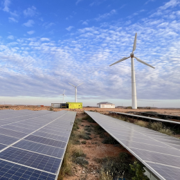 A solar panel field in the foreground with wind turbines and buildings in the background. There are clouds floating above. The sky is bright blue.