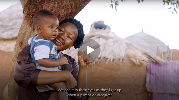 Video screenshot of a woman holding her baby while smiling and pointing. The child is mesmerized by what she is pointing at off-screen.