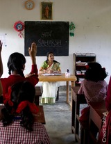Students eagerly raise their hands to answer a question and a teacher is seated facing them, smiling. 