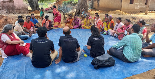 A group of adults sit outside on a blue mat in discussion with three individuals with shirts that say “Locally Led Development Growing Together.”