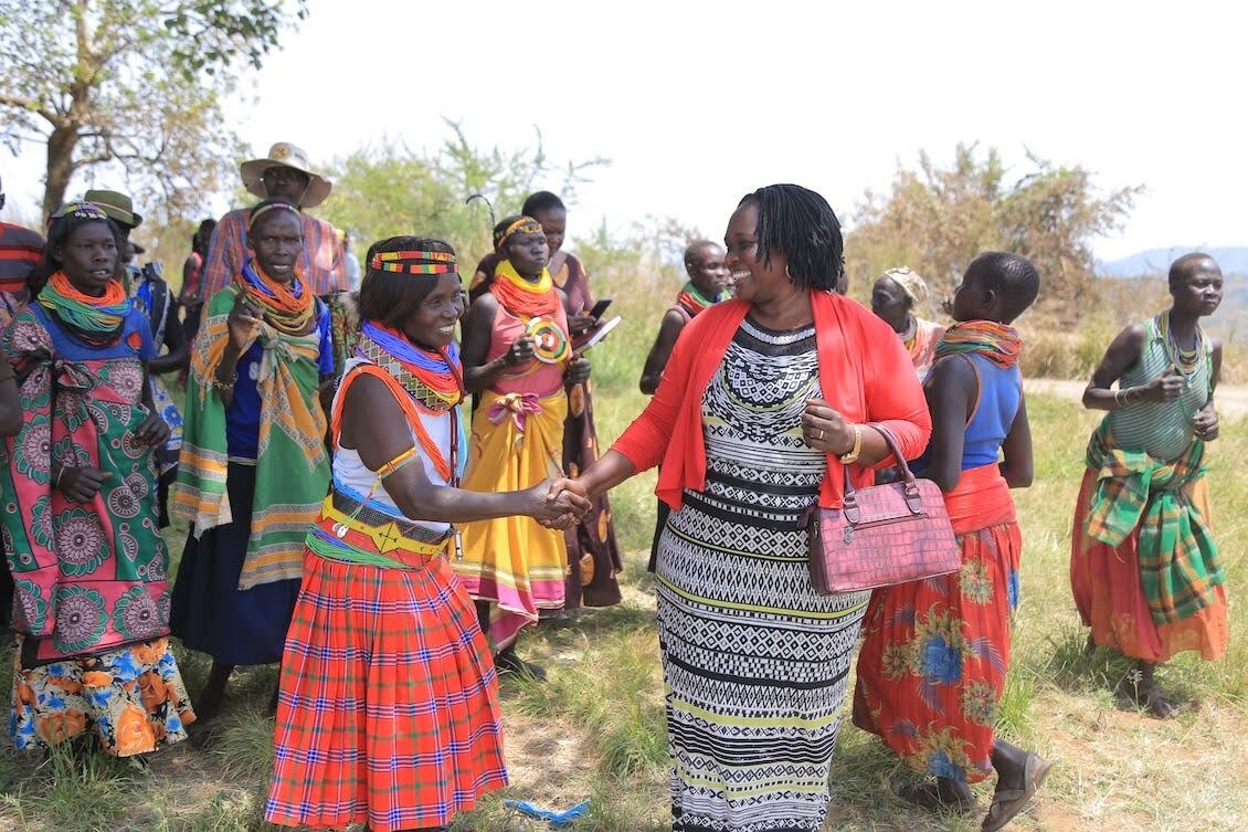 Two women shake hands smiling at one another, surrounded by members of an indigenous community in an outdoor setting in Uganda.