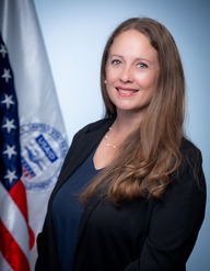 A woman with long brown hair, wearing a suit, smiles for a photo; US and USAID flags are visible in the background