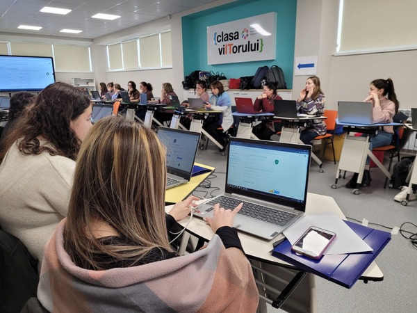 About 20 women sit at desks facing each other in a room with a screen at one end. In the foreground, a woman facing a laptop is typing on her machine.