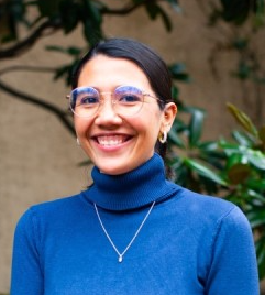 Photo of a smiling woman wearing a blue turtleneck and glasses with dark hair in a bun.