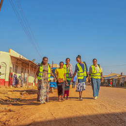Four women in neon yellow safety vests walk along a dirt road with another young woman in a yellow shirt