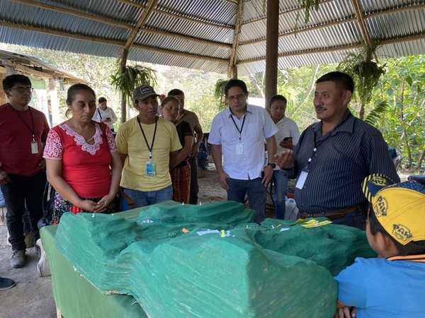 People gathering outdoors around a table with a green model design of a valley looking at a man in a navy blue striped shirt talking. 