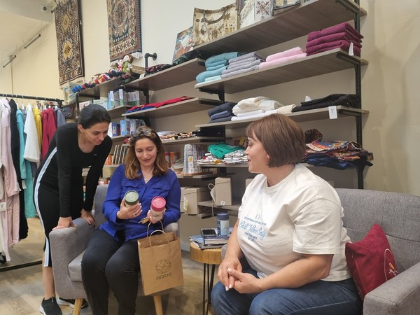 3 women in a store admire goods held by 1 woman. 2 women sit in gray chairs in front of shelves. 1 woman stands, leaning over to look at products.