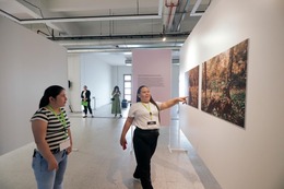  Woman points at one of 2 large photographs hanging on a wall at an exhibit, while another woman observes from further back.