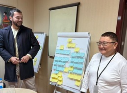 2 individuals stand indoors on opposite sides of a flip chart covered in yellow and blue sticky notes with writing on them.