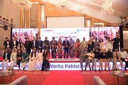 A group of 32 people stand and sit on a stage, pointing to a sign that reads “Justuju” in Urdu, followed by “Under Local Works Pakistan” in English.