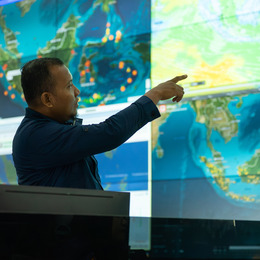 A man in a dark long sleeve shirt points at a wall of screens filled with weather maps