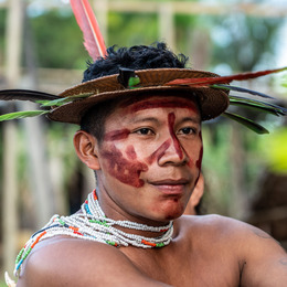 An indigenous man wearing a straw headdress with feather and white bead necklace. His face is painted with red markings.