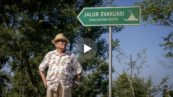 A man in a floral shirt stands underneath a green sign with "JALUR EVAKUASI Evacuation route" and a volcano on it