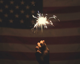 Photograph of a hand holding a sparkler in front of an American flag. 