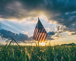 Photograph of a small American flag sitting in a field of grass with the sunrising behind it.