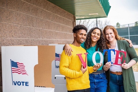 Three adults stand outside a polling location urging passers-by to vote.