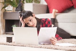 A young woman looks at her laptop while laying down on her living room floor.