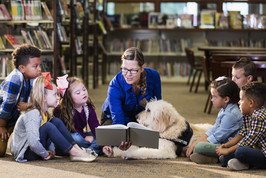 A woman, accompanied by a therapy dog, sits on the floor of a library and reads to six children. 