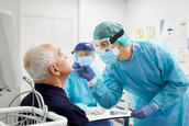 A physician wearing PPE administers a nasal swab test to an elderly patient, while another physician wearing PPE looks on in the background.