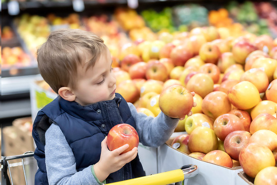 A child sitting in a grocery cart pics out apples from the produce section of a supermarket.
