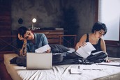 Two women file through paperwork while sitting on a bed.