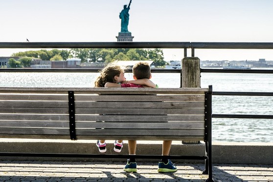 Two kids sit on a bench at a scenic viewpoint that looks out at the Statue of Liberty.