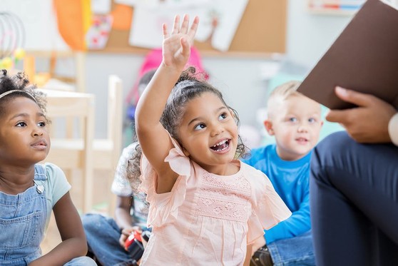 A preschool aged girl raises her hand to be called upon in class.