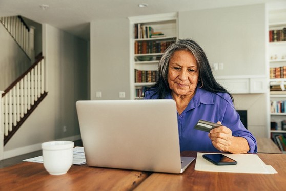 An older woman sits at a table with her laptop, phone, and credit card.
