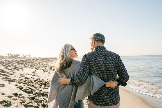 An older couple walk along a beach while holding hands.