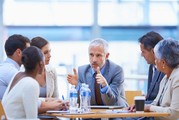 A man speaks to a group of men and women at a table about business matters.