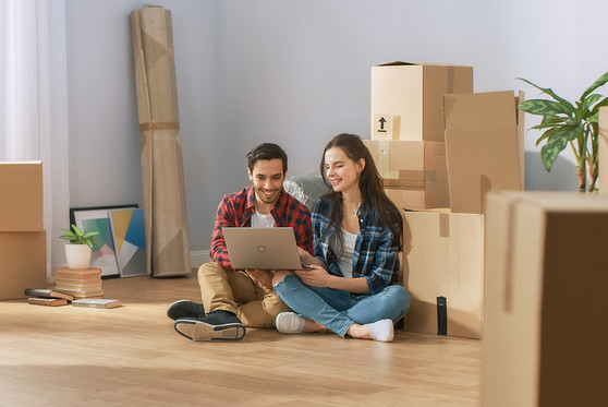 A man and woman sit on the floor of their new home using their laptop while leaning against boxes..