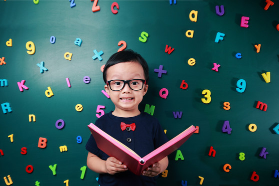 A child of Asian descent holds an open book and stands in front of a green background with various colored letters and numbers.